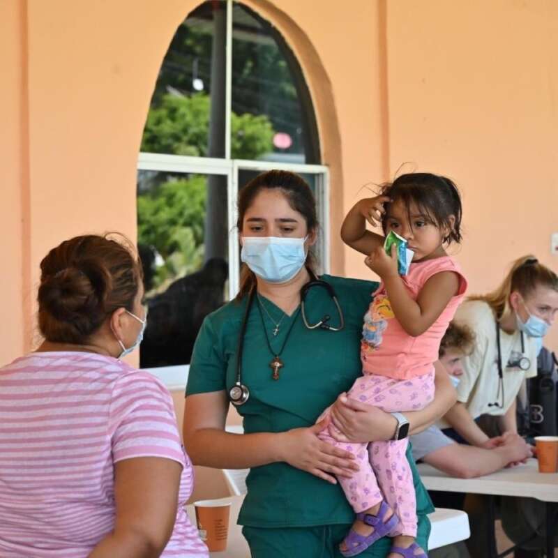 A masked health worker holds a child in her arms while speaking to another person