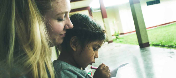A student looks over a young girl's shoulder as she writes on a piece of paper