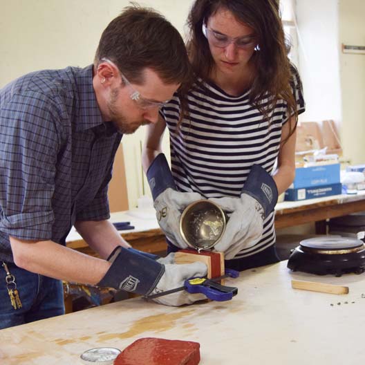 Prof. Bryan Park assists a student in pouring molten metal into a cast for Benedictine College's Discovery Scholar Medal