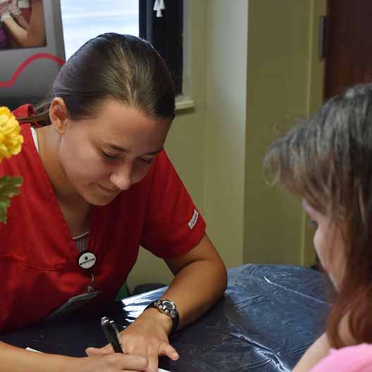 A nursing student writes on a piece of paper across a desk from a woman